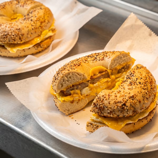 Two everything bagels with sausage, egg, and melted cheese on white paper plates.