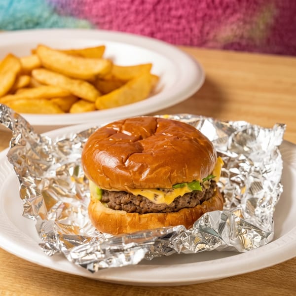 A cheeseburger wrapped in foil on a white plate, with a plate of fries in the background.